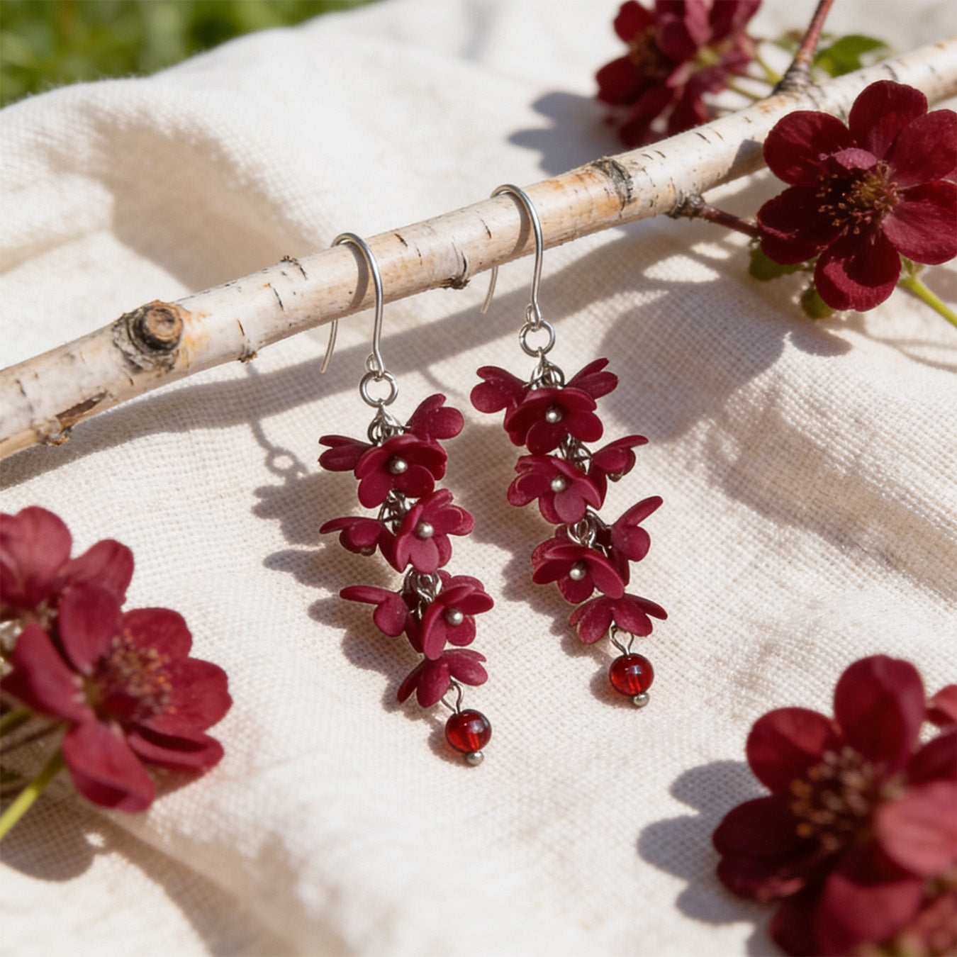 Shadow Vine Burgundy Earrings displayed on a birch branch among burgundy flowers in bright natural daylight. Handmade in Finland byMinta.