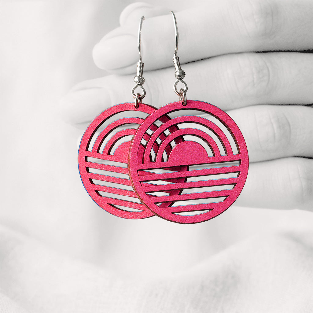 Raspberry Aamu (Sunrise) plywood earrings made in Finland, featuring a geometric sunrise design and silver-toned hooks, photographed hanging from a hand against a softly blurred light linen background.