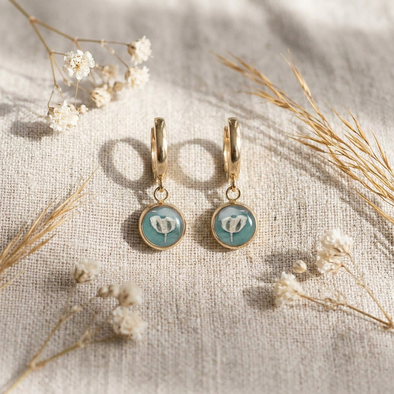 Delicate sage green botanical huggie earrings styled on linen with dried flowers and warm natural light