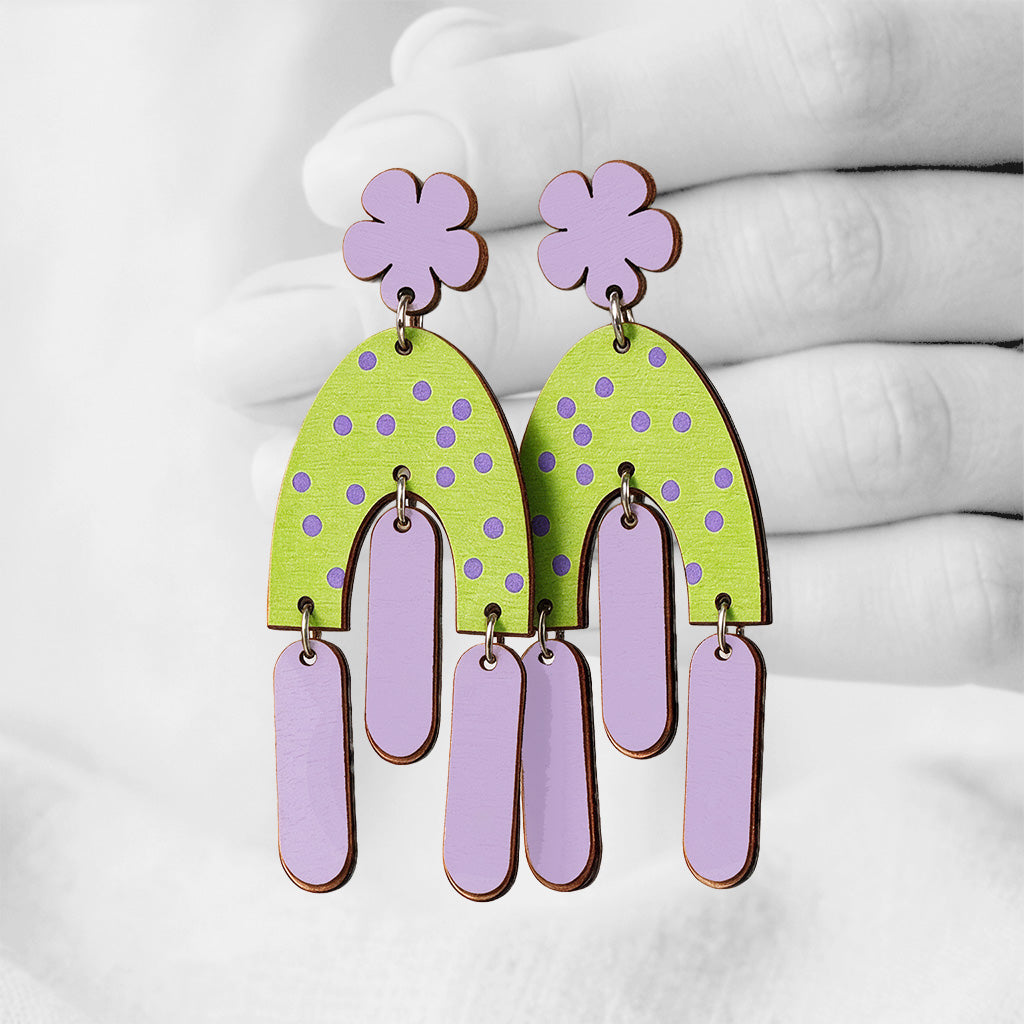 Purple and green Helmat Heiluen (Swinging Hems) plywood stud-earrings made in Finland, photographed against a black and white background of a hand.
