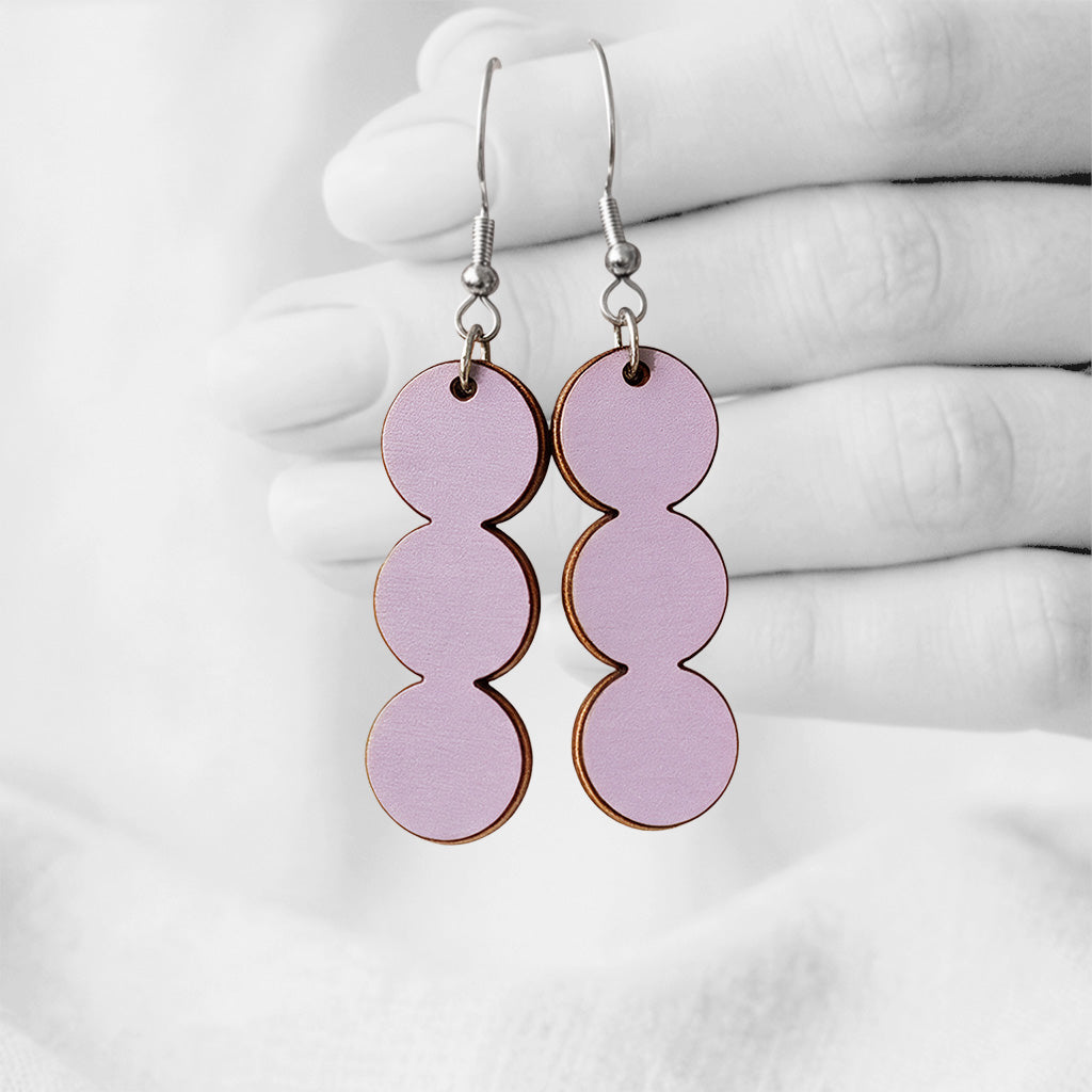 Smoke Lavender String of Pearls plywood earrings made in Finland, photographed hanging from a hand against a softly blurred light linen background.