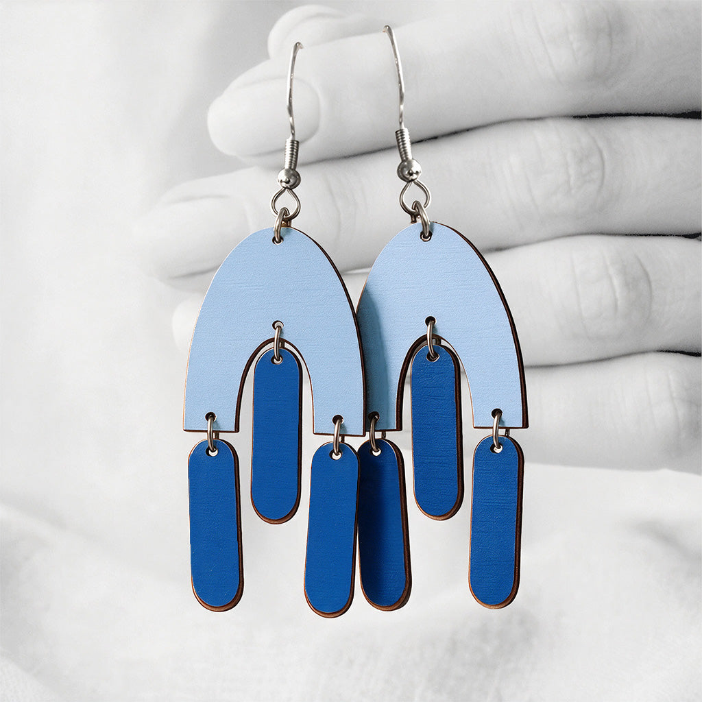 Blue and light blue HulmuHelma (Fluttering Hems) plywood earrings made in Finland, photographed against a black and white background of a hand.