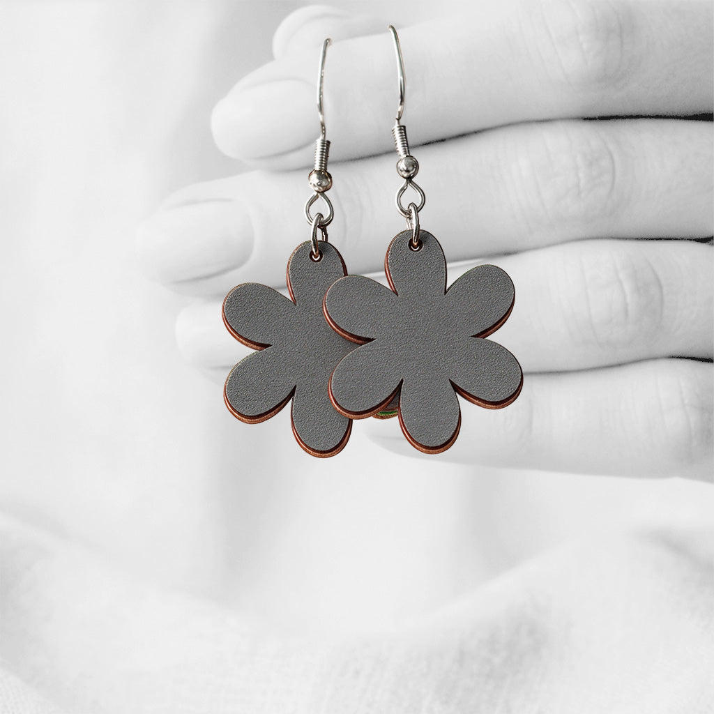 Grey Kakkara flower-shaped earrings made from Finnish birch plywood, photographed hanging from a hand in black and white against a softly blurred light background.