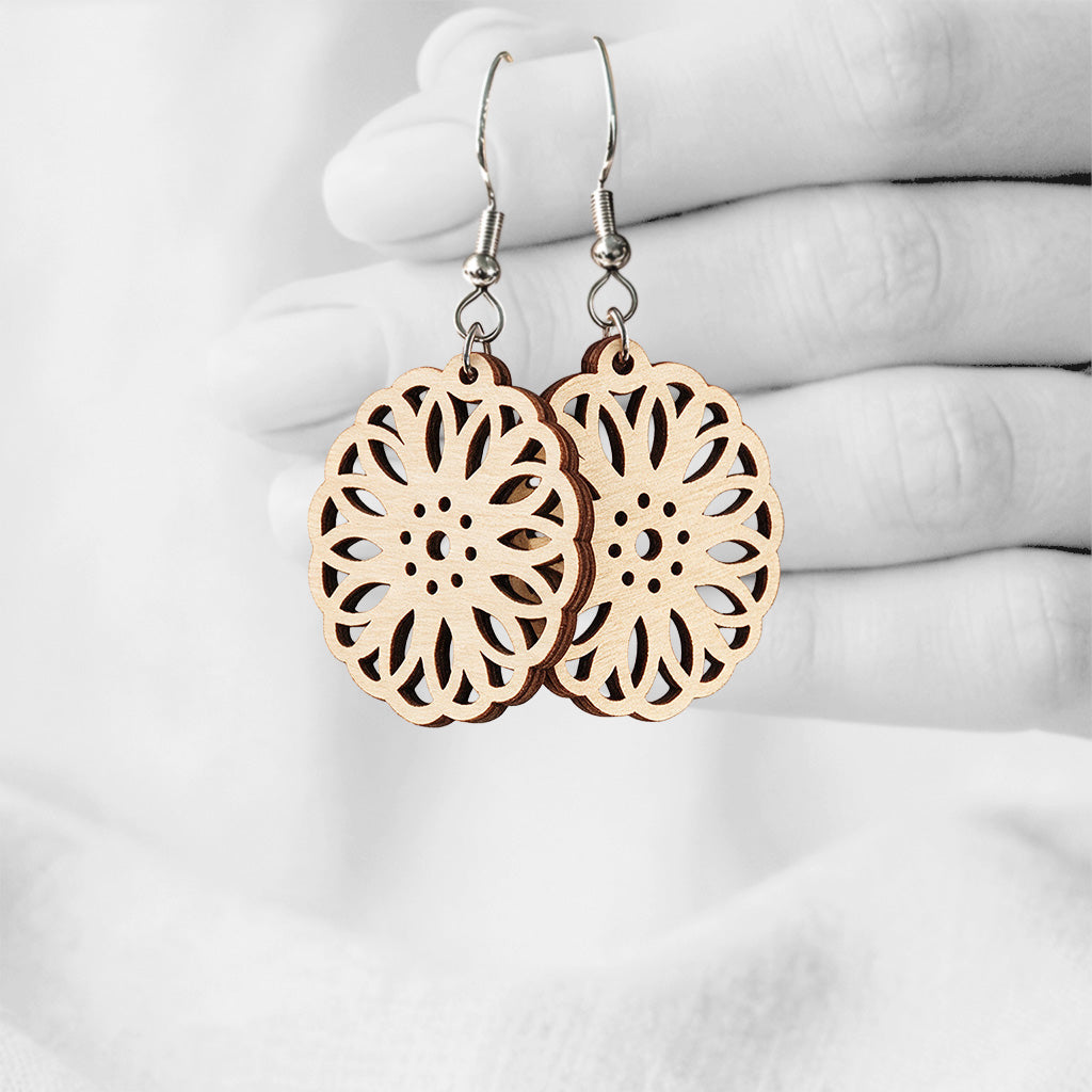 Natural wood Kaleidoskooppi plywood earrings with intricate floral laser-cut details, made in Finland, photographed hanging from a hand in black and white against a softly blurred light background.