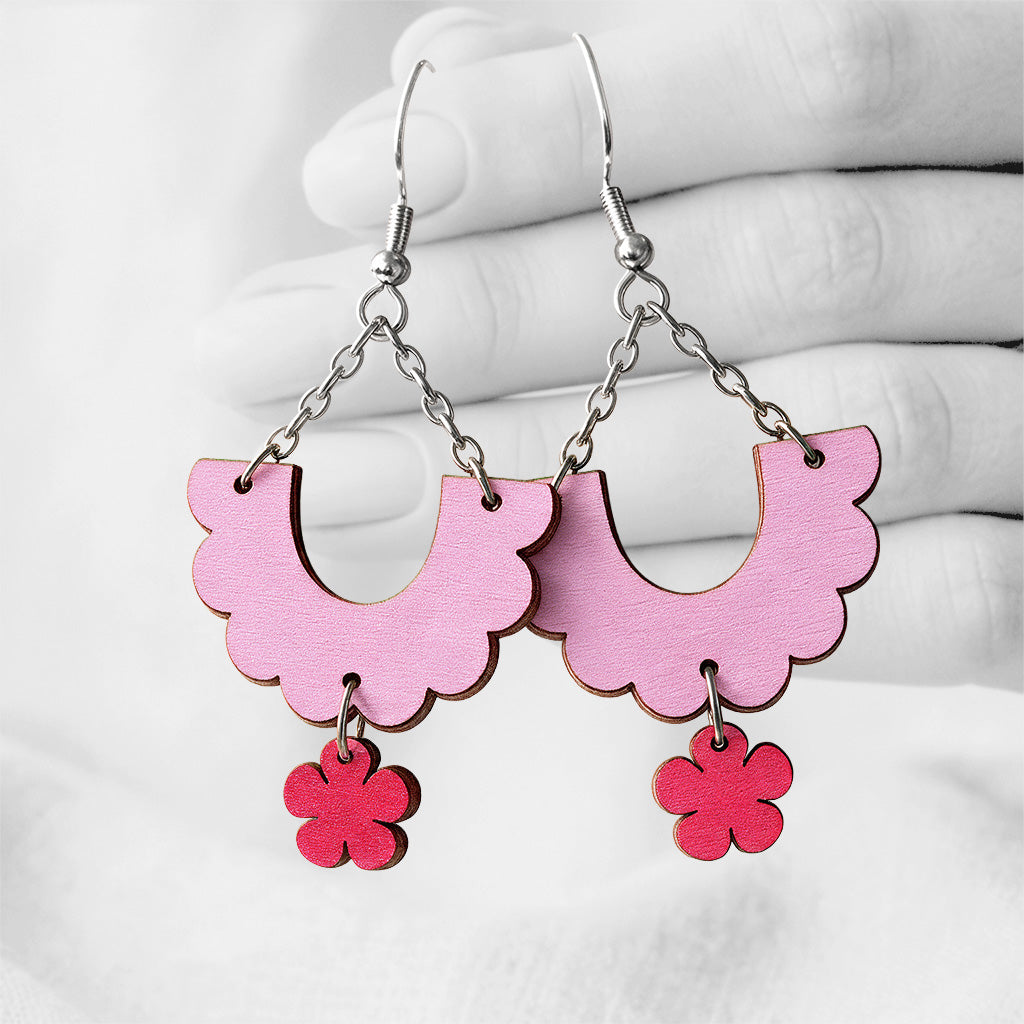 Malva and raspberry Kiikku (Swing) plywood earrings handmade in Finland, photographed hanging against a black-and-white hand background.