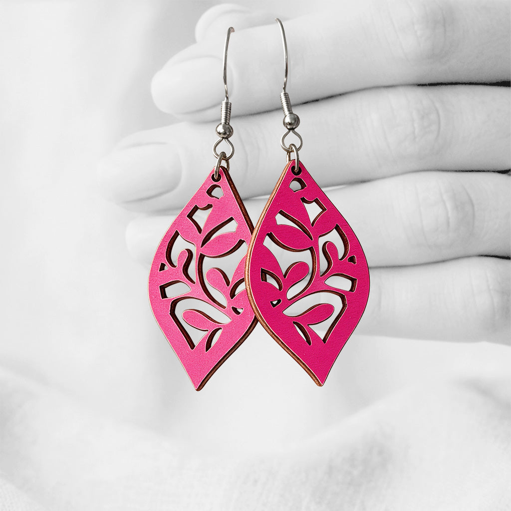 Raspberry Köynnös plywood earrings with intricate floral cut-out details, made in Finland, photographed hanging from a hand in black and white against a softly blurred light background.
