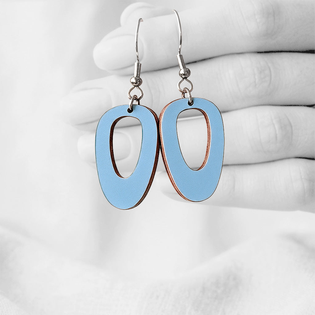 Light blue Luoto (Islet) plywood earrings made in Finland, photographed hanging from a hand in black and white against a softly blurred light background.