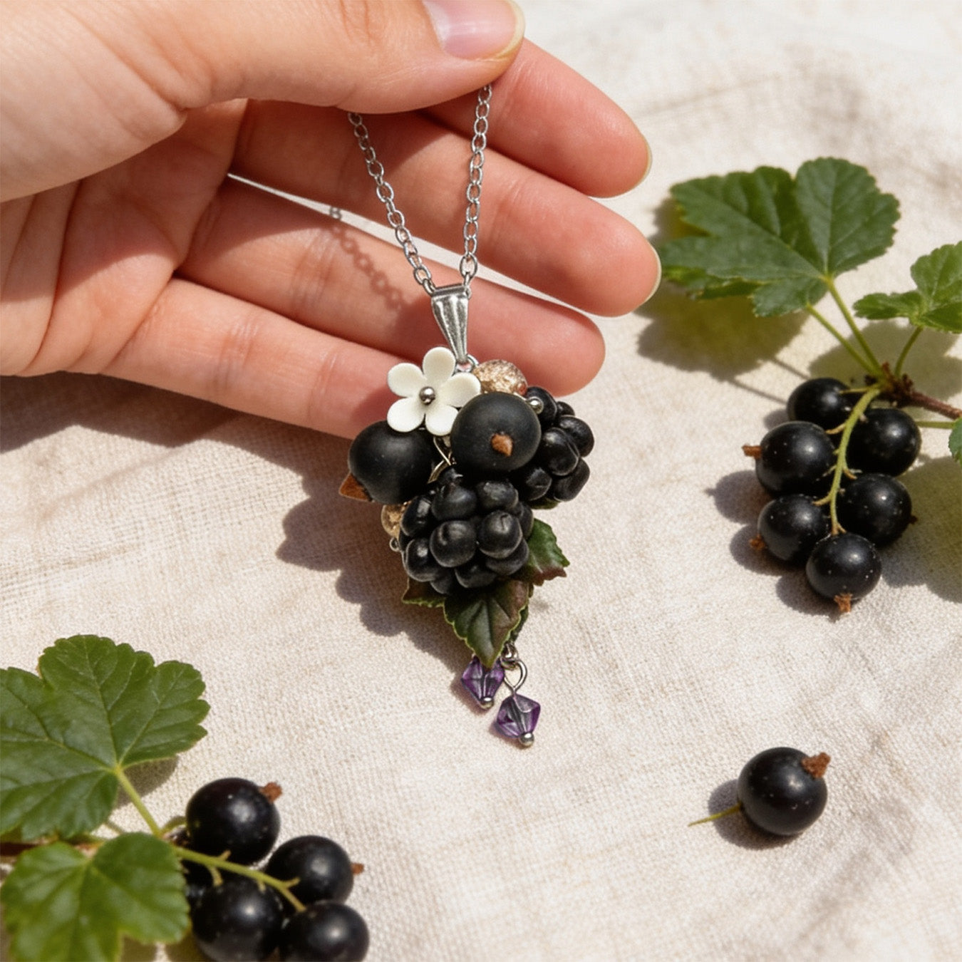 Hand holding a handmade berry necklace for size reference, featuring sculpted blackcurrants, blackberries, detailed green leaves, a small white flower and purple crystal accents. Surrounded by real black currants and currant leaves on soft linen in bright natural daylight.