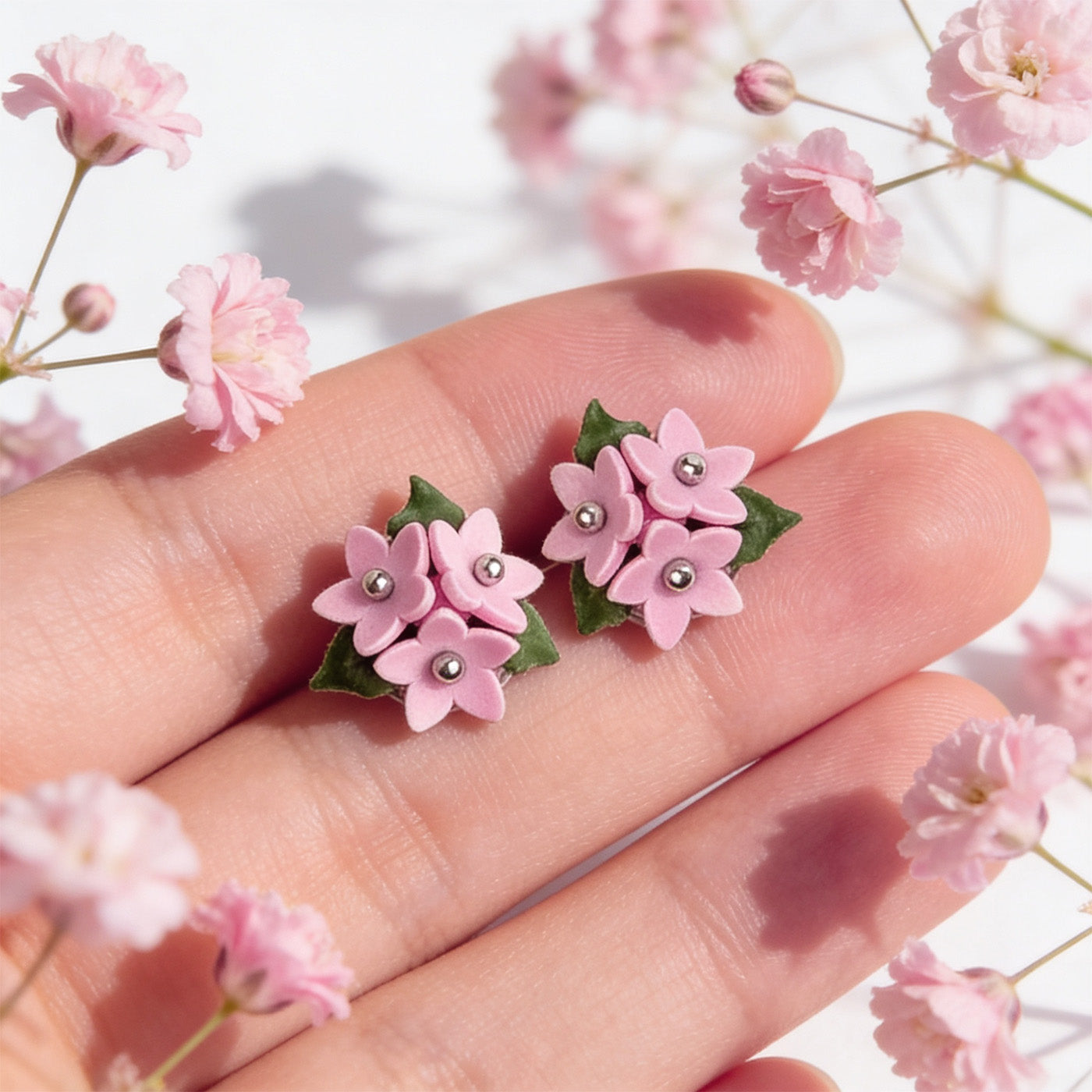 Pink Forest Star stud earrings handmade by byMinta resting on fingertips among soft pink flowers in bright natural daylight.