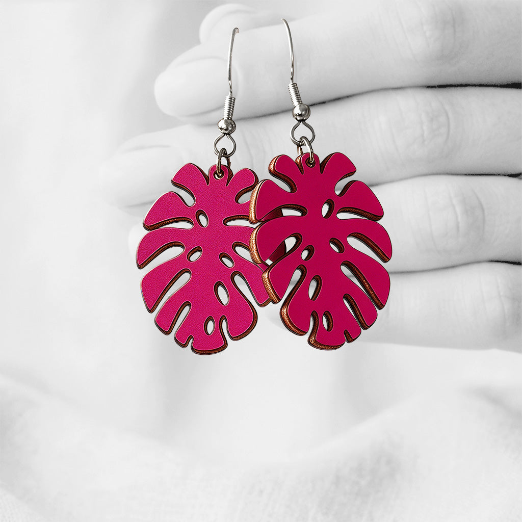 Raspberry Monstera plywood earrings made in Finland, photographed hanging from a hand in black and white against a softly blurred light background.