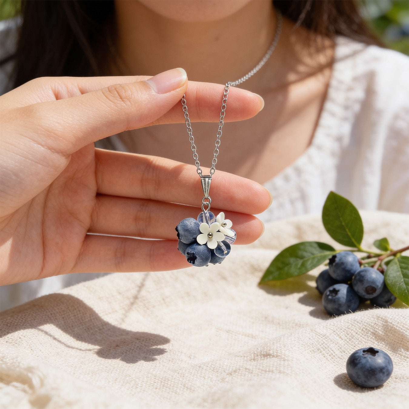 Blueberry Bouquet Necklace held gently in hand for scale, featuring blueberries and delicate flowers. Natural linen fabric, fresh blueberries, and green leaves in soft natural light. Handmade in Finland by byMinta.