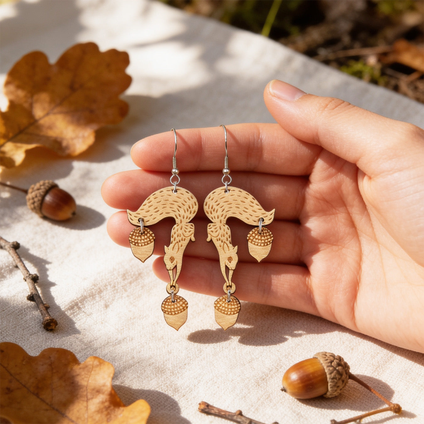 Hand holding handmade squirrel earrings for size reference among oak leaves, acorns and small twigs. Lightweight Finnish birch plywood earrings photographed in warm natural sunlight with a soft Nordic autumn forest atmosphere.