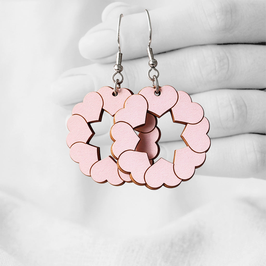Light pink Pusu (Kiss) plywood earrings made in Finland, photographed hanging from a hand in black and white against a softly blurred light background.