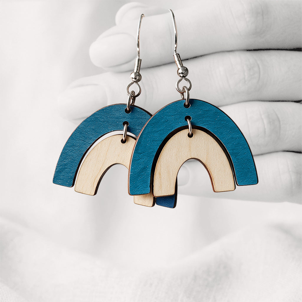 Bluepetrol and birch Riemukaari Duo plywood earrings handmade in Finland, shown against a softly blurred hand and light linen background