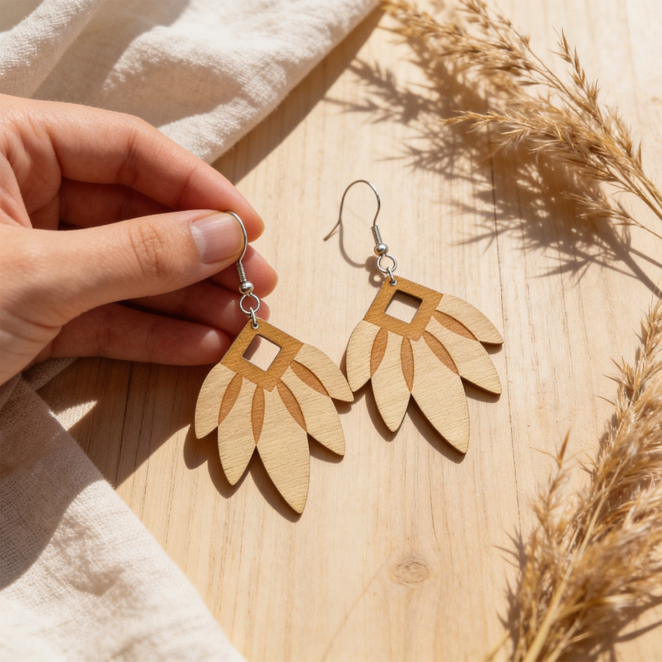 Hand holding handmade wooden leaf earrings for size reference on a light wooden surface. Warm natural sunlight, soft linen fabric and dried grasses create an airy Nordic atmosphere with natural styling.