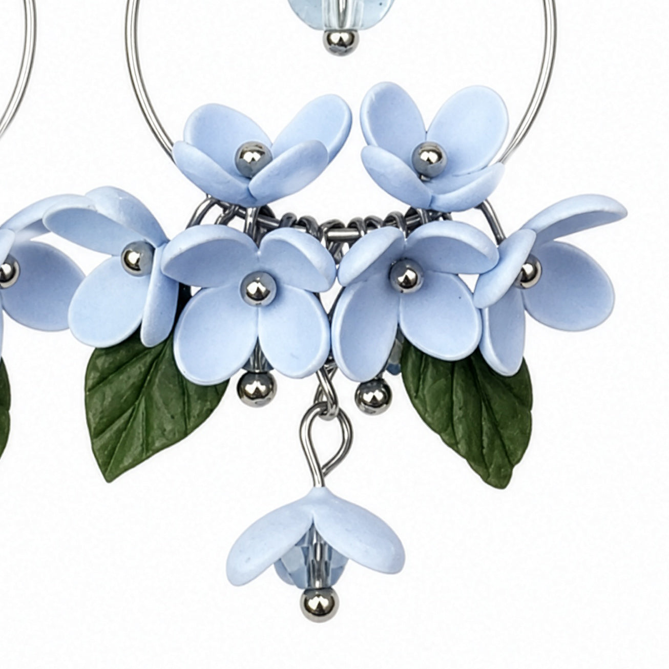 Close-up detail of Mist Flower Hoop Earrings showing pale blue blossoms, green leaves, and handcrafted floral elements. Handmade in Finland by byMinta.