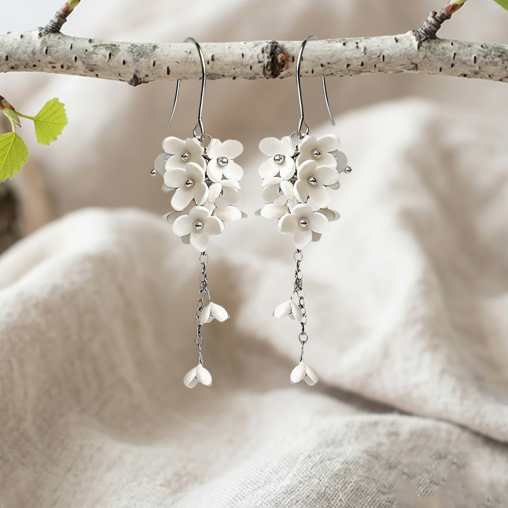 Handcrafted white Jasmike flower earrings hanging from a birch branch on natural linen fabric, made in Finland.