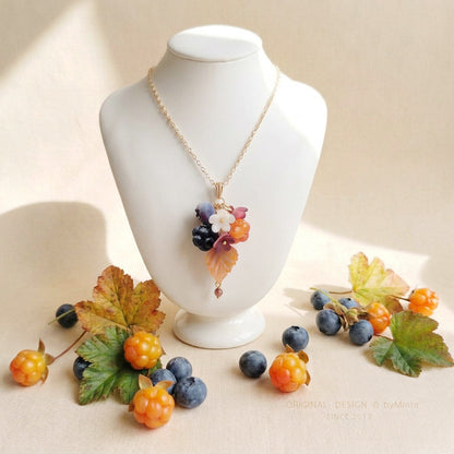 Marjametsä necklace with a gold-plated chain, photographed on a white jewelry display among berries and autumn leaves in natural light