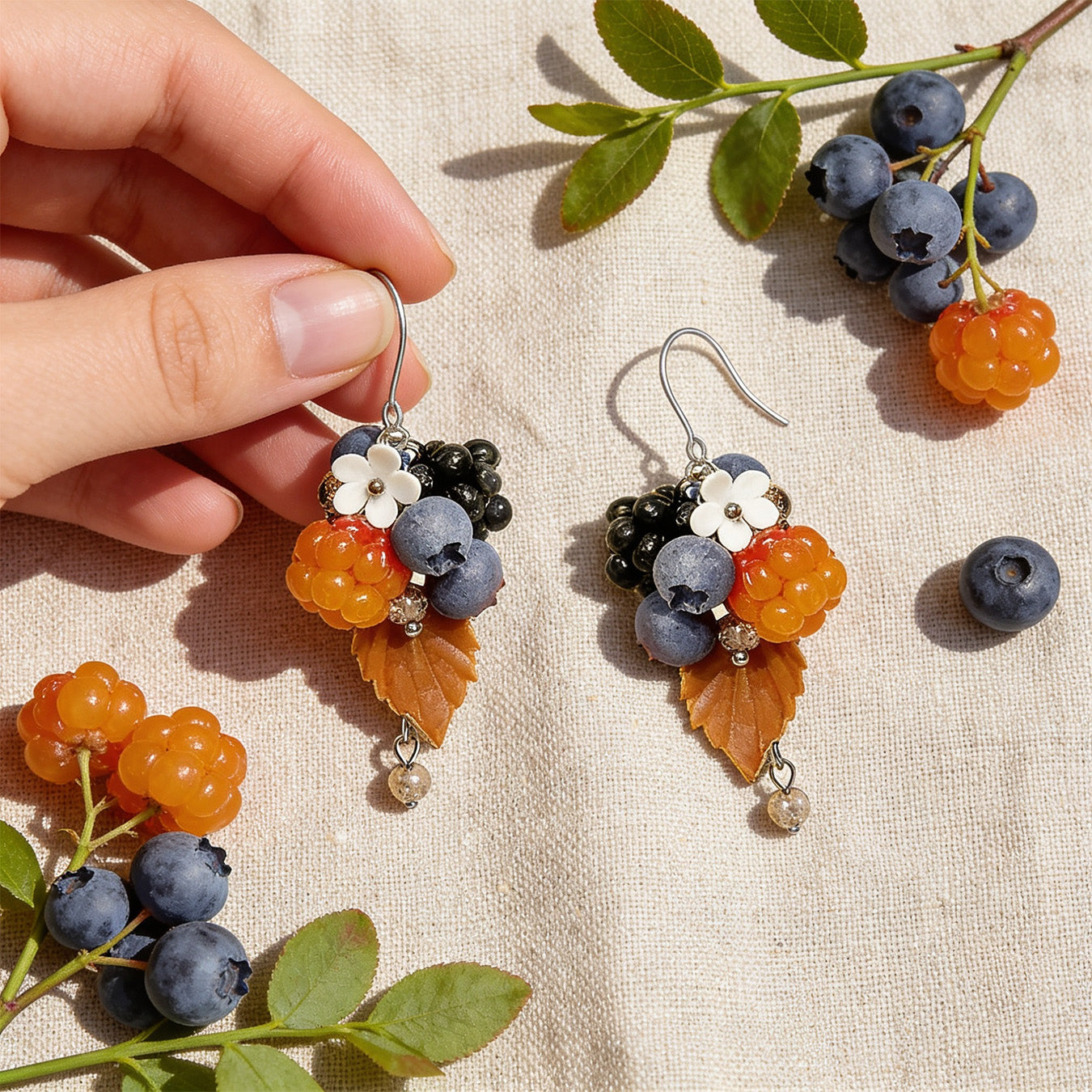 Berry Forest Earrings photographed with fresh cloudberries, blueberries, and green leaves in soft natural light, with one earring gently held by hand for scale. Handmade in Finland by byMinta.