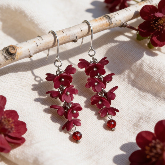 Shadow Vine Burgundy Earrings displayed on a birch branch among burgundy flowers in bright natural daylight. Handmade in Finland byMinta.
