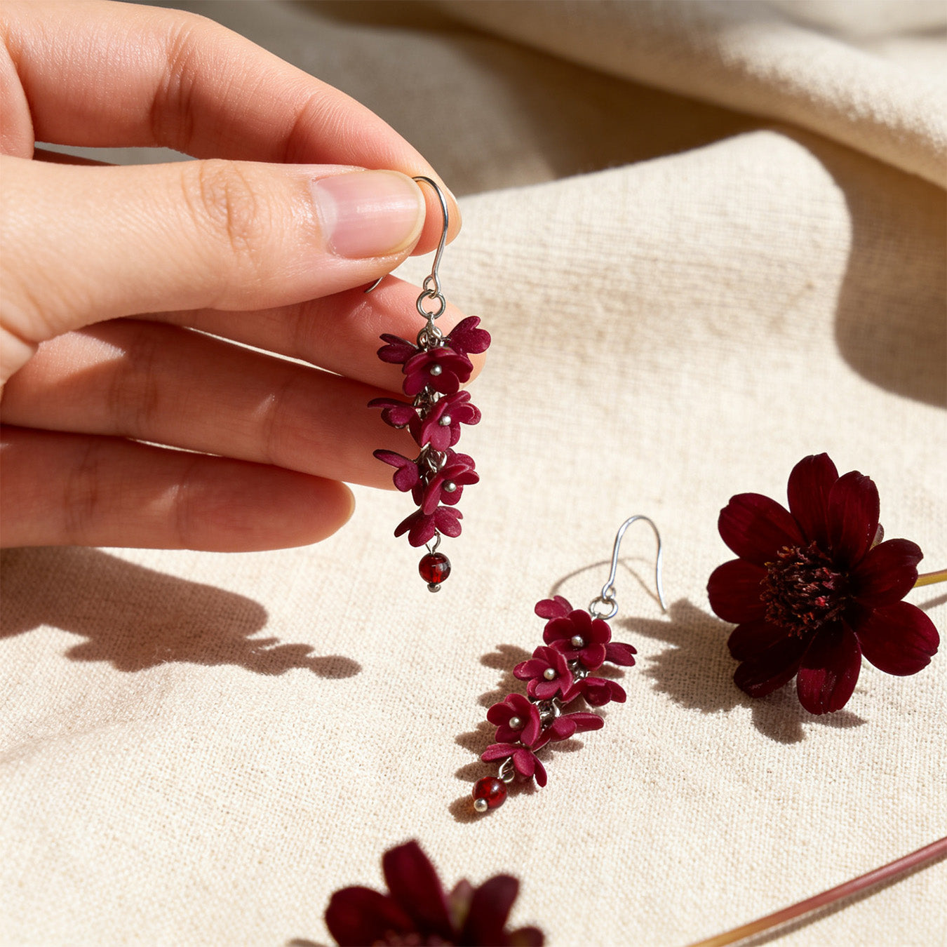 Shadow Vine Burgundy Earrings photographed with deep burgundy flowers in soft natural light, with one earring gently held by hand for scale. Handmade in Finland by byMinta.