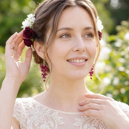 Bride in a sunlit summer garden wearing delicate burgundy floral earrings by byMinta. Her hair is styled in a soft updo adorned with deep burgundy roses and small white blossoms, and she wears an elegant ivory lace wedding dress. She smiles gently while adjusting a flower in her hair, creating a romantic and timeless bridal look inspired by natural beauty and handcrafted details.