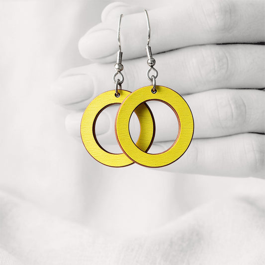Yellow Circle plywood earrings made in Finland, photographed hanging from a hand in black and white against a softly blurred light background.