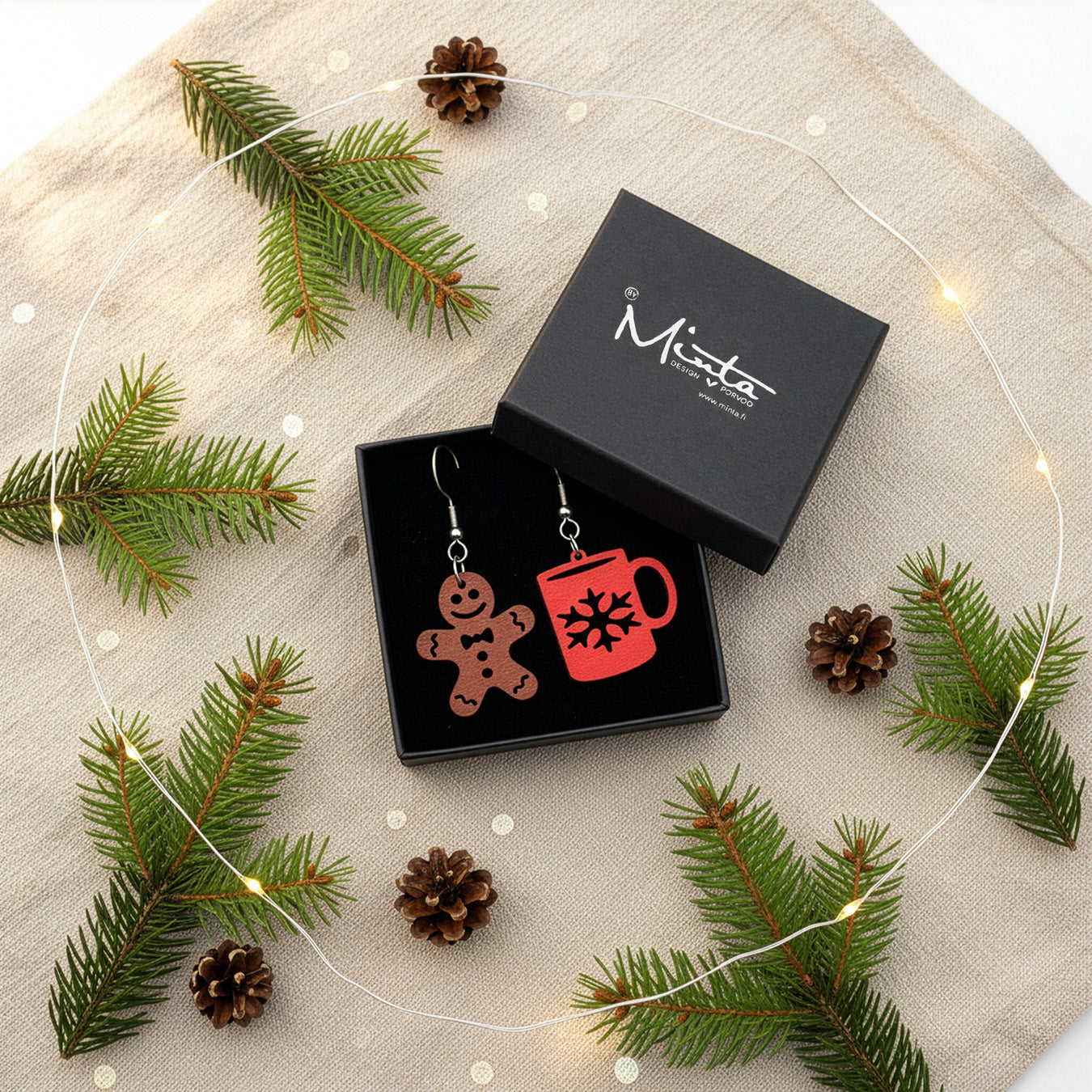 Gingerbread cookie and mug earrings presented in a gift box with pine branches, pinecones, and warm fairy lights, creating a cozy festive atmosphere. Handmade in Finland by byMinta.