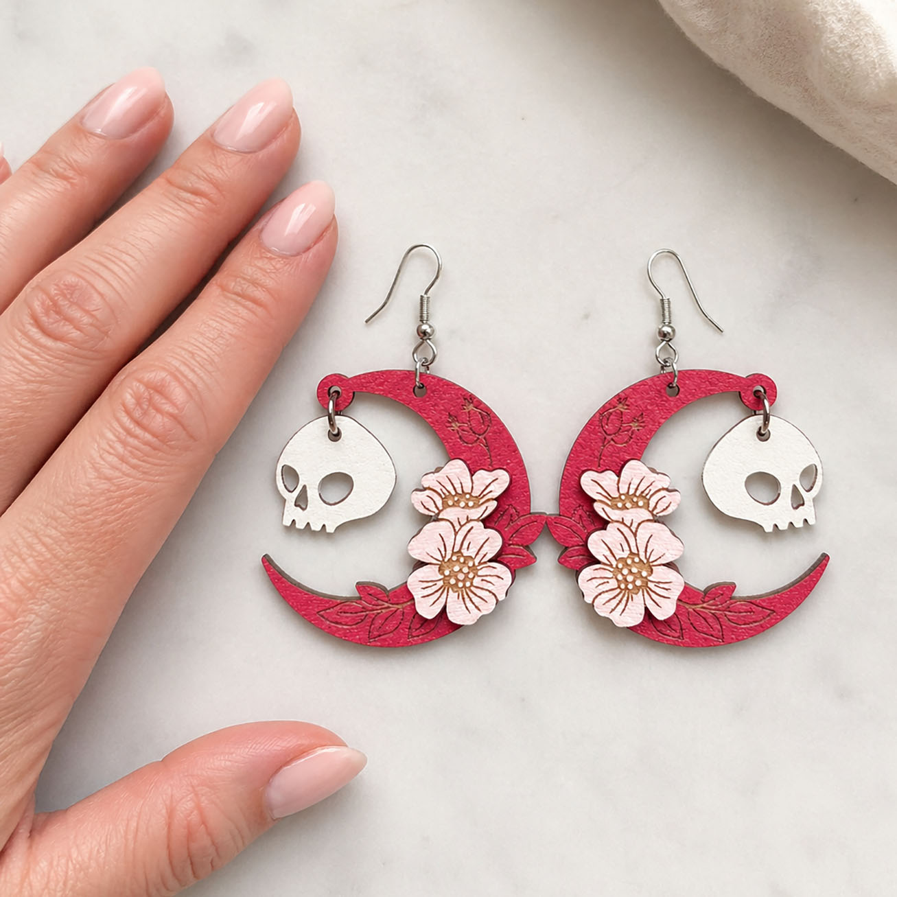 Handmade pink crescent moon skull earrings with flowers Finland byMinta, lightweight wooden jewelry, shown with hand for scale