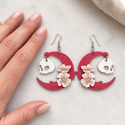 Handmade pink crescent moon skull earrings with flowers Finland byMinta, lightweight wooden jewelry, shown with hand for scale