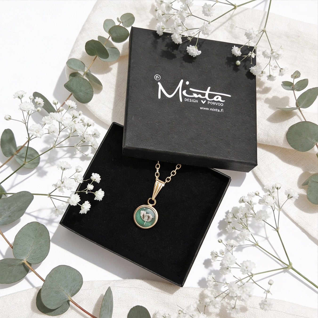 Handmade botanical necklace displayed in a black jewelry box surrounded by delicate baby's breath flowers and soft green eucalyptus leaves. Elegant product photography in bright natural daylight with a light linen background.