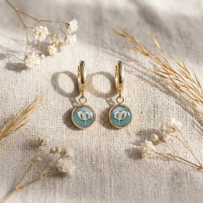 Delicate sage green botanical huggie earrings styled on linen with dried flowers and warm natural light