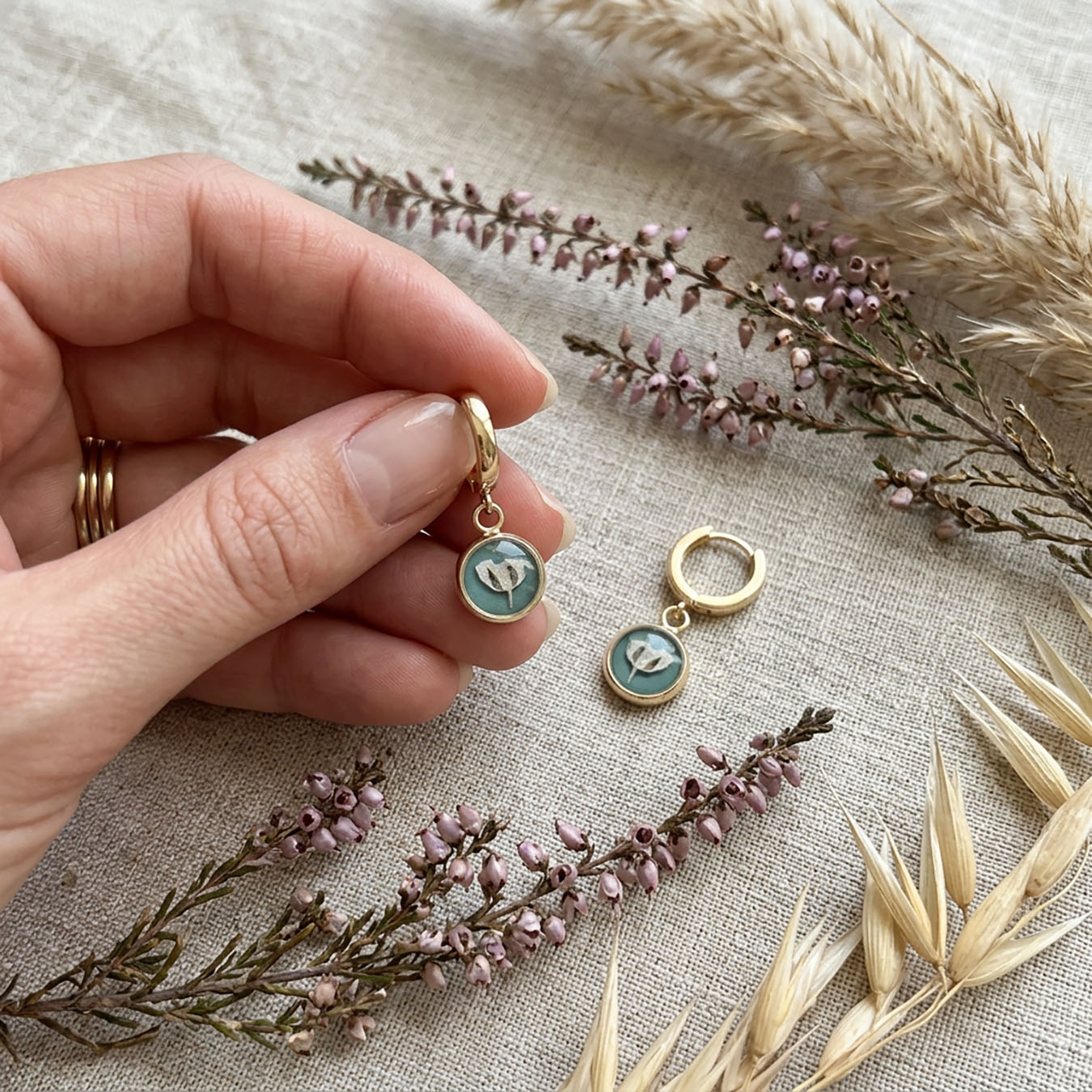 Hand holding sage green floral huggie earring on linen fabric with heather and dried grasses