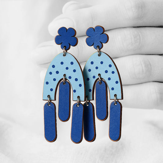 Blue Helmat Heiluen (Swinging Hems) plywood stud-earrings made in Finland, photographed against a black and white background of a hand.