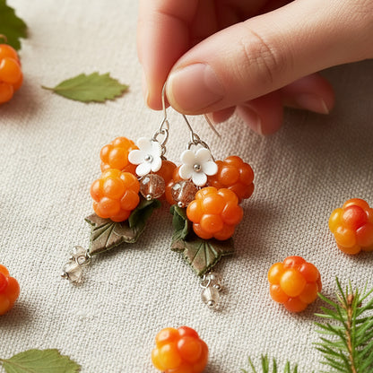 Cloudberry Marsh Earrings held gently in hand for scale, surrounded by natural linen fabric, fresh cloudberries, and green leaves. Handmade in Finland by byMinta.