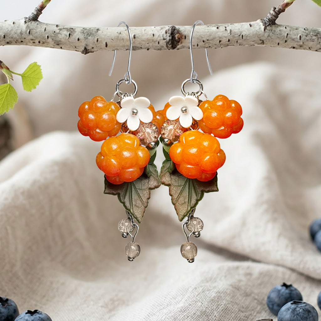 Handcrafted cloudberry earrings hanging from a birch branch, featuring blueberries, flowers and green leaves.