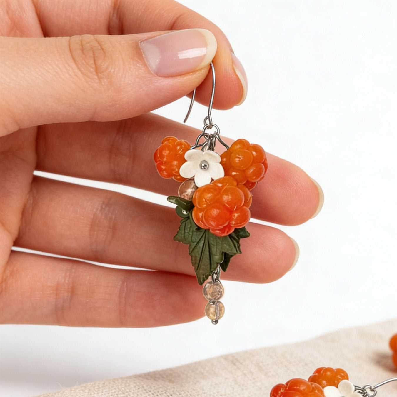 Cloudberry Marsh Earrings held gently in hand for scale, surrounded by natural linen fabric, fresh cloudberries, and green leaves. Handmade in Finland by byMinta.