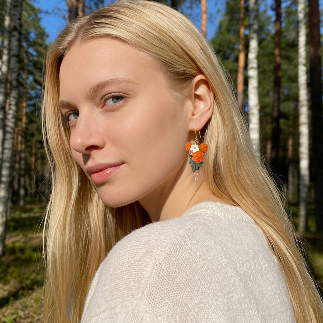 Blonde woman wearing Cloudberry Marsh Earrings in a sunlit Nordic forest. Handmade earrings inspired by the warm glow of cloudberries.