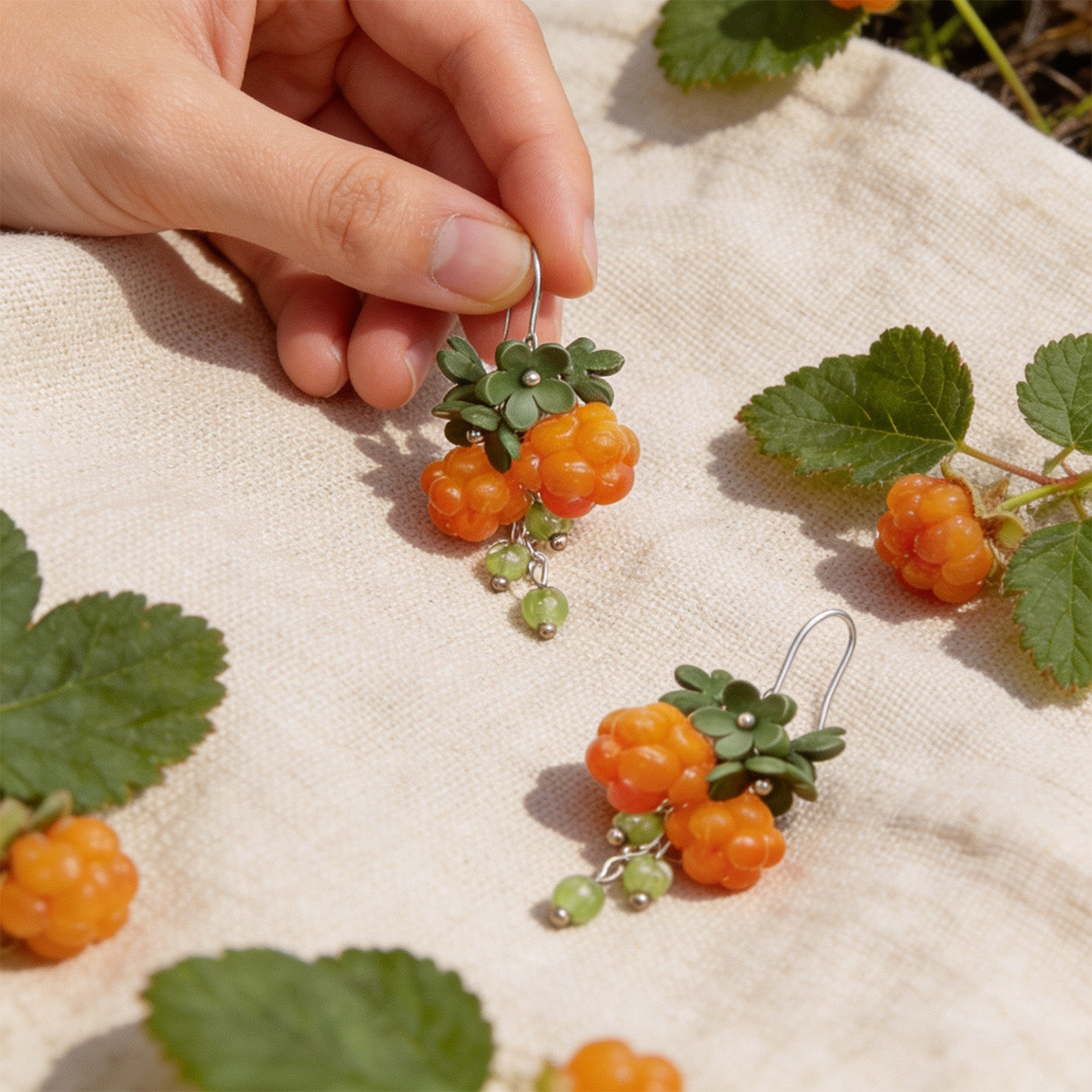 Cloudberry Sprout Earrings photographed with fresh cloudberries and green leaves in soft natural light, with one earring gently held by hand for scale. Handmade in Finland by byMinta.