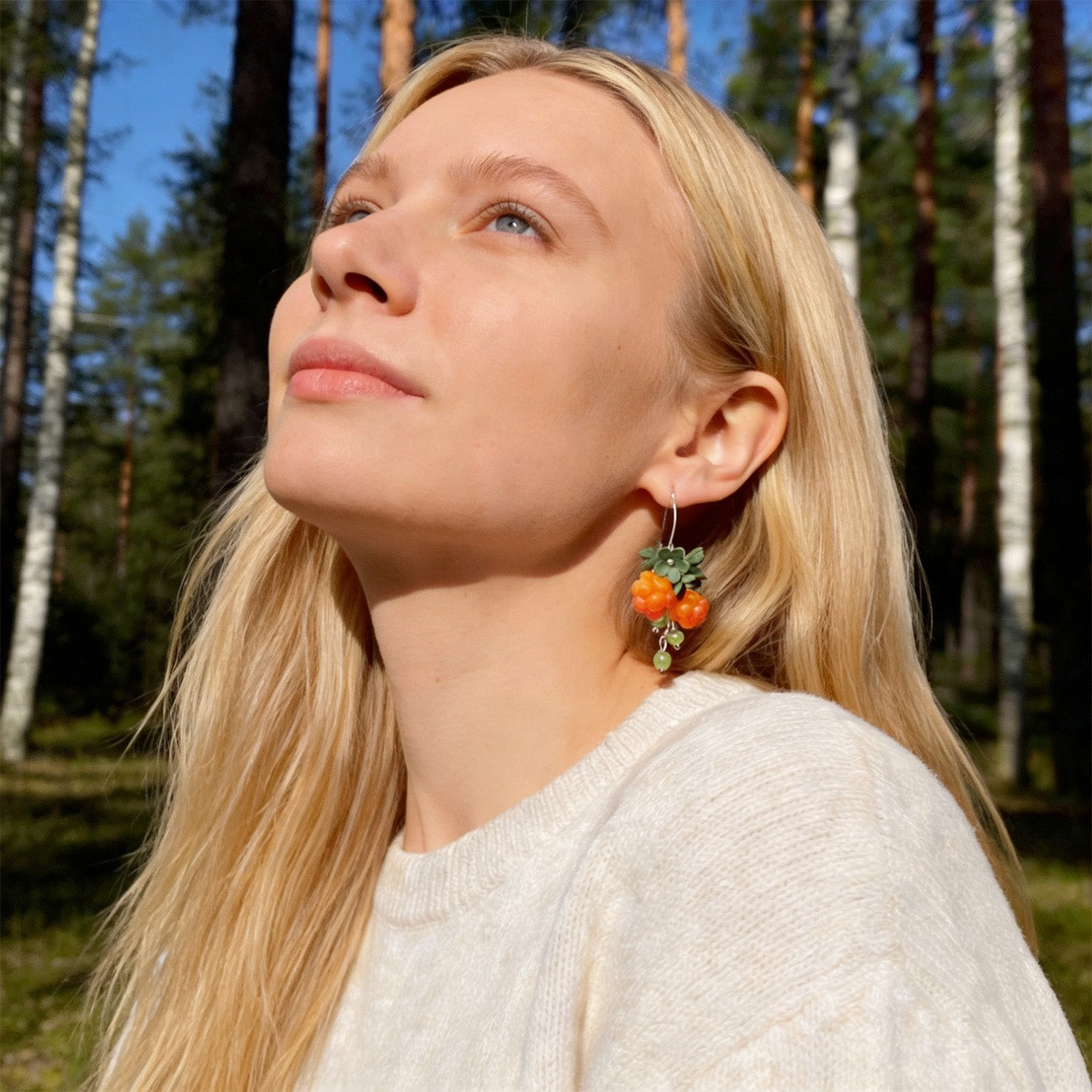 Blonde woman wearing Cloudberry Sprout Earrings in a sunlit Nordic forest. Handmade earrings inspired by the warm glow of ripe cloudberries.