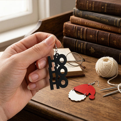 Hand holding handmade HO HO HO and Santa Claus earrings on a wooden table with vintage books, knitting yarn, and reading glasses, creating a cozy Christmas atmosphere. Handmade in Finland by byMinta.