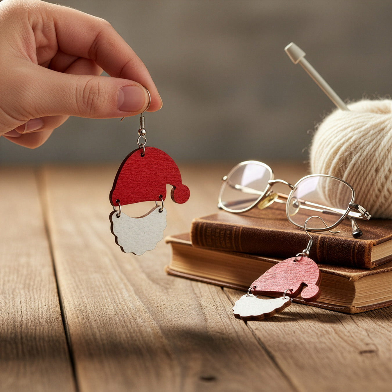 Hand holding a Santa Claus earring on a wooden table with books, glasses, and yarn, creating a cozy holiday atmosphere. Handmade in Finland by byMinta.