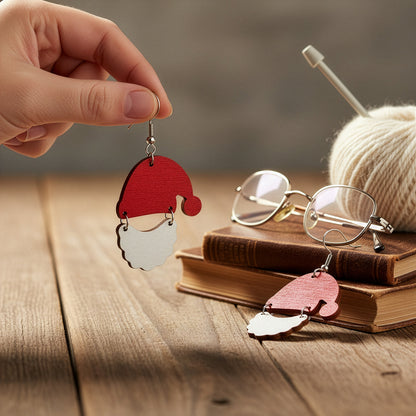 Hand holding a Santa Claus earring on a wooden table with books, glasses, and yarn, creating a cozy holiday atmosphere. Handmade in Finland by byMinta.