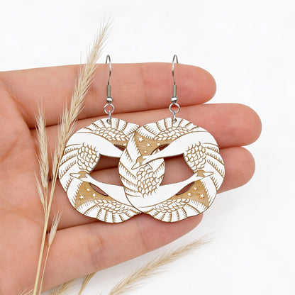 Wooden swan earrings by byMinta with detailed engraved feather patterns, photographed on a hand for scale with soft natural reed stems.