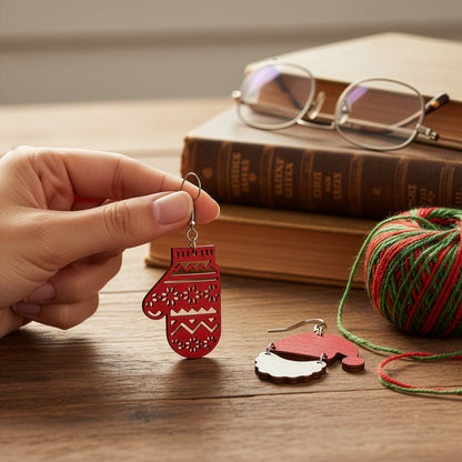 Hand holding the red mitten earring on a wooden table with books, yarn, and glasses, creating a cozy Christmas atmosphere. Handmade in Finland by byMinta.