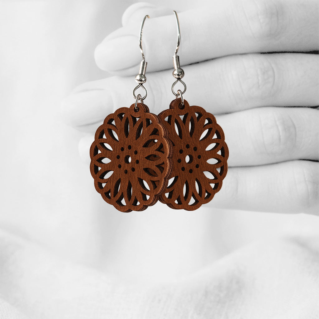 Walnut Kaleidoskooppi plywood earrings with intricate floral laser-cut details, made in Finland, photographed hanging from a hand in black and white against a softly blurred light background.