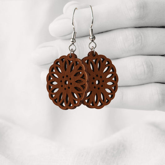 Walnut Kaleidoskooppi plywood earrings with intricate floral laser-cut details, made in Finland, photographed hanging from a hand in black and white against a softly blurred light background.