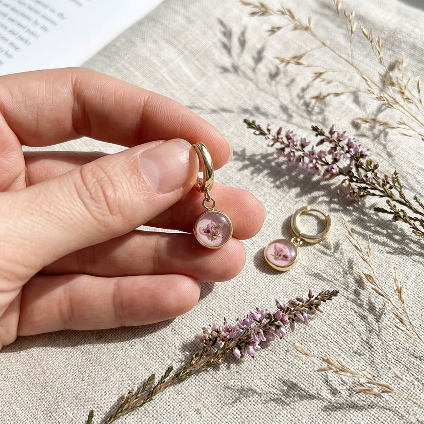 Hand holding pink heather flower huggie earring with real flower detail on soft linen background