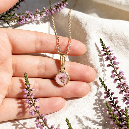 Hand holding a handmade botanical necklace for size reference, featuring a real heather flower on a soft dusty pink background. Surrounded by delicate purple heather blooms and linen in bright natural daylight with a soft Nordic autumn atmosphere.