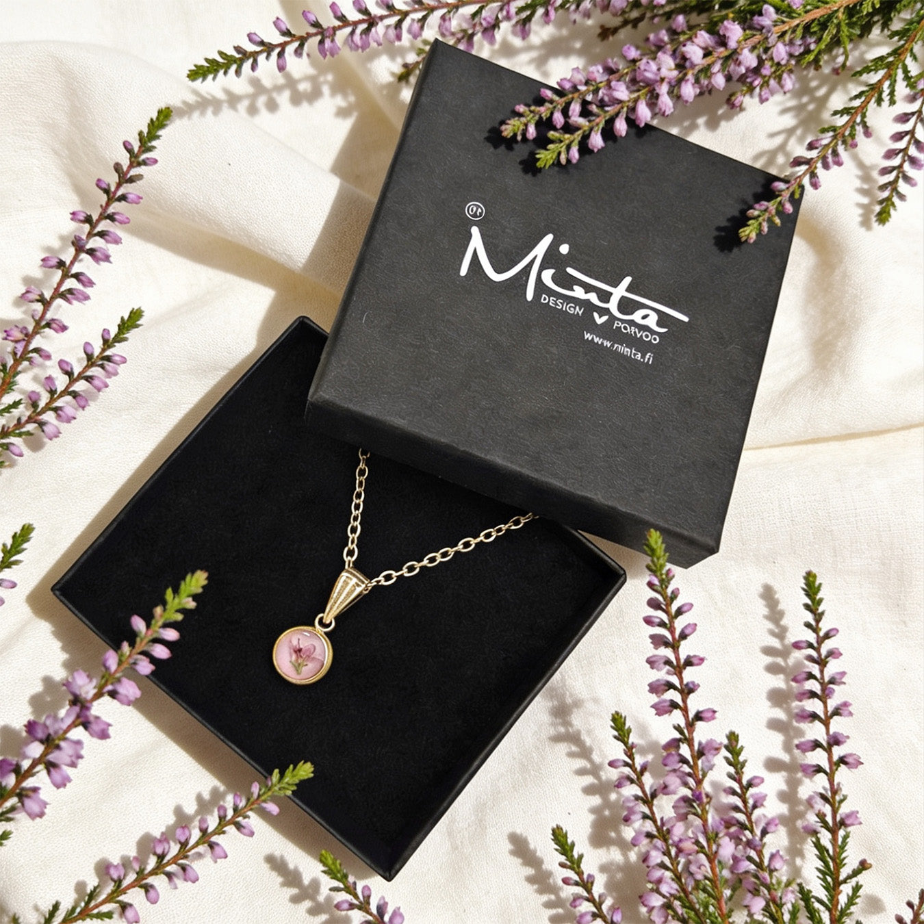 Handmade botanical necklace with a real heather flower displayed in a black byMinta jewelry box. Surrounded by delicate purple heather blooms and soft linen in bright natural daylight with a gentle Nordic autumn atmosphere.